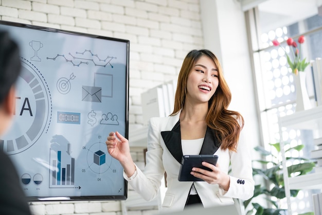 A cheerful and confident Asian businesswoman stands and uses a tablet to present a bar chart of data from a monitor to her office colleagues Focus on Asian business women leader role at the meeting