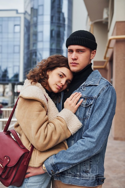 Cheerful couple in casual warm clothes embracing each other outdoors in the city near business building