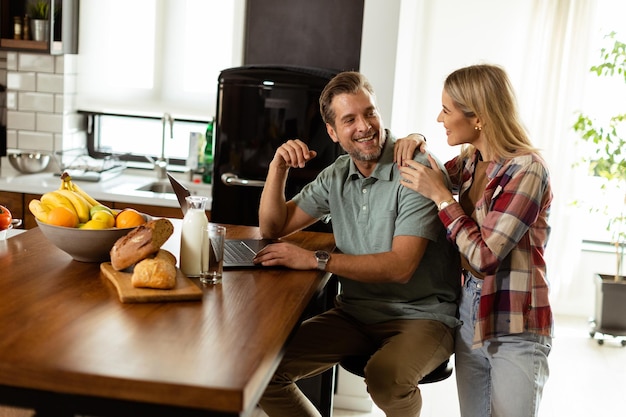 Cheerful couple enjoys a lighthearted moment in their sunny kitchen working on laptop surrounded by a healthy breakfast