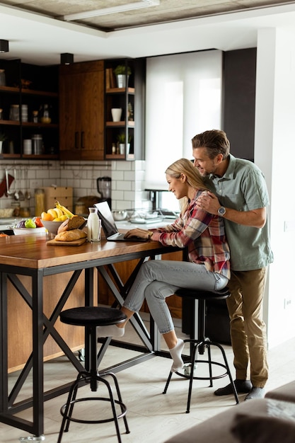 Cheerful couple enjoys a lighthearted moment in their sunny kitchen working on laptop surrounded by a healthy breakfast