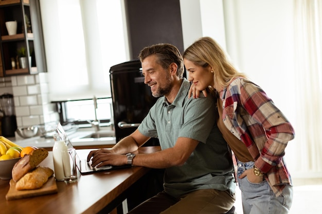 Cheerful couple enjoys a lighthearted moment in their sunny kitchen working on laptop surrounded by a healthy breakfast