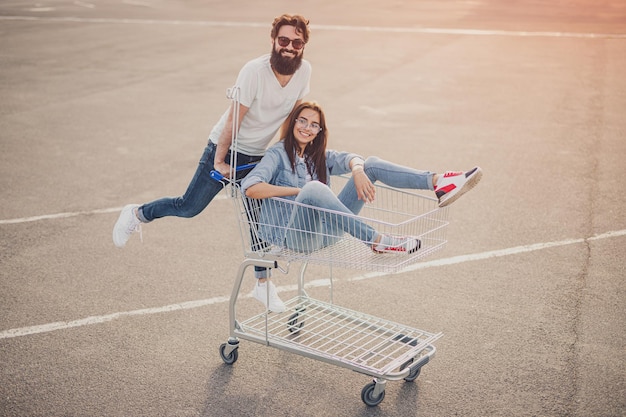 Cheerful couple riding shopping cart on parking