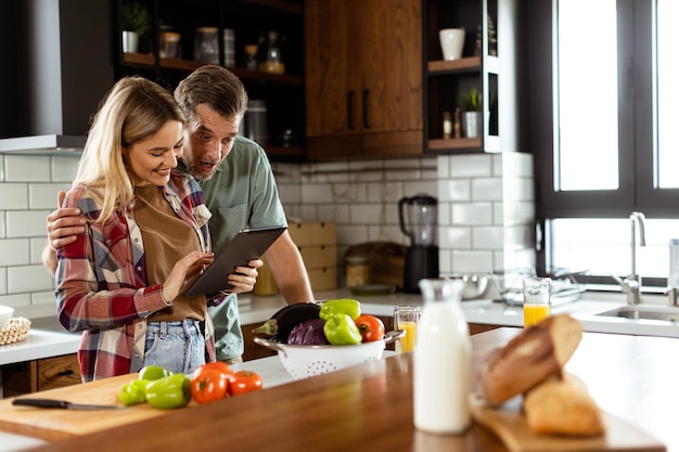 Cheerful couple stands in a welllit kitchen engrossed in a digital tablet among fresh ingredients