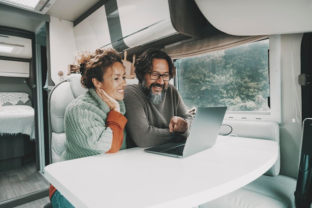 Cheerful couple using laptop together inside a camper van alternative house Man and woman surfing the web or working on computer sitting and relaxing in indoor leisure activity during rv travel