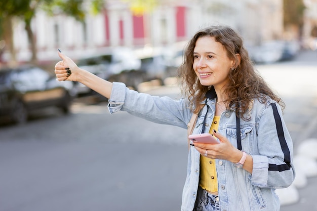 Cheerful curly hair woman in denim jacket catches car in the city at bright sunny day