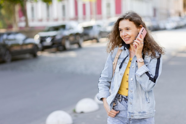 Cheerful curly hair woman in denim jacket talking on phone at bright sunny day