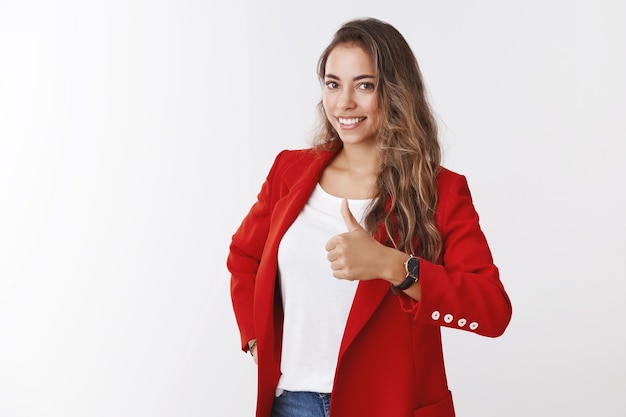 Cheerful curly-haired caucasian woman white teeth smiling wearing red trendy jacket showing thumb up like approval gesture, grinning approvingly, congratulating friend great choice, good work