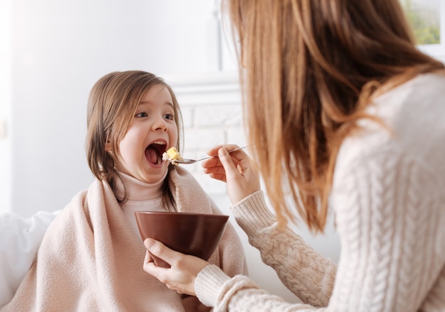 Cheerful cute little girl eating breafast with her loving mother while sitting together on the couch and resting at home