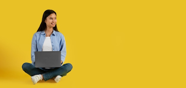 Cheerful cute millennial korean lady student in casual sit on floor typing on computer look at blank space