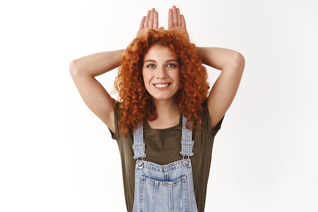 Cheerful cute redhead curlyhaired woman with blue eyes having fun enjoy weekends looking playful and smile positive showing rabbit ears with hands above head standing white background