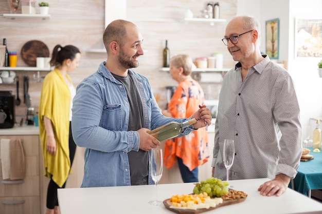 Cheerful dad and son talking in kitchen while the rest of the family is busy in meal preparation. Man holding bottle of wine.