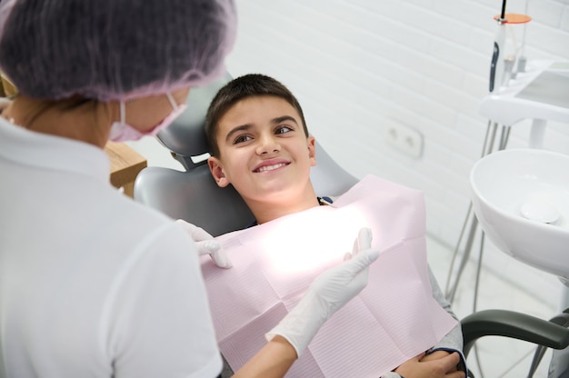 Cheerful dark-haired schoolboy lying on dentist's chair, smiles with beautiful toothy smile looking at dental assistant preparing him for regular medical check-up. Dentistry, oral care hygiene concept