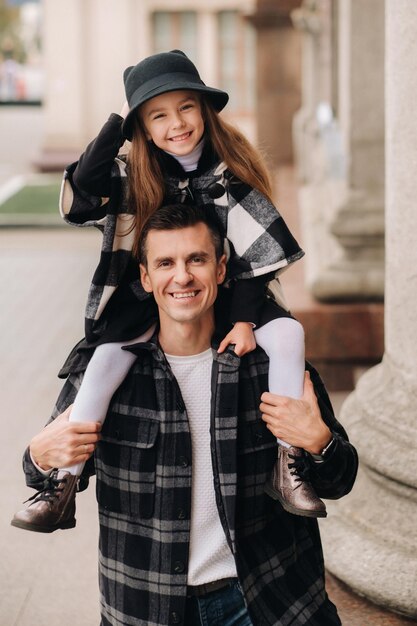 A cheerful Daughter is sitting on dad's shoulders and walking around the autumn city