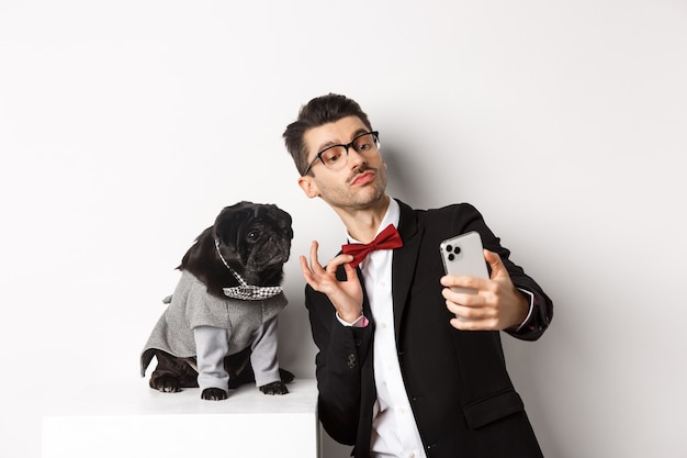 Cheerful dog owner in suit celebrating New Year with dog, taking selfie on smartphone near cute black pug in costume, white.