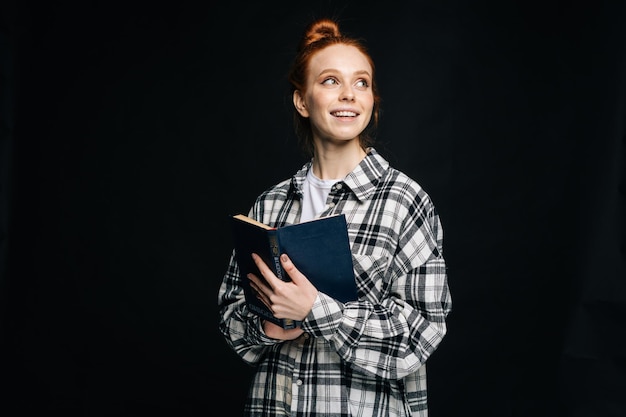 Cheerful dreamy young woman college student holding opened book and looking away on isolated black background Pretty redhead lady model emotionally showing facial expressions