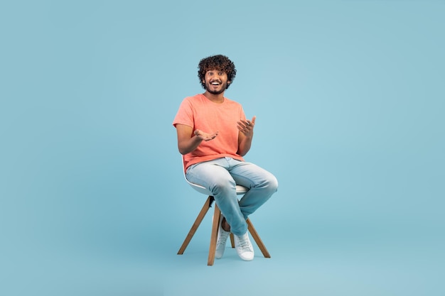 Cheerful eastern guy sitting on chair over blue background