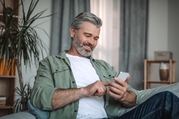 Cheerful elderly european man with beard typing on smartphone enjoying social media chat in room interior