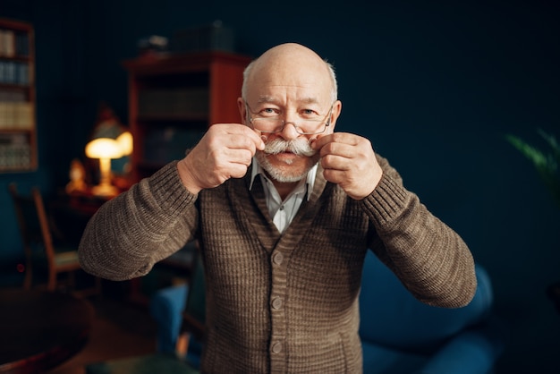 Cheerful elderly man shows his moustache in home office. Bearded mature senior poses in living room, old age businessman