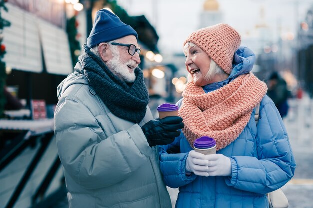 Cheerful elderly woman holding a carton cup of coffee and laughing while being outdoors on sunny winter day with her husband