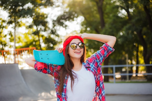 Cheerful emotional young woman dressed in stylish clothes while holds skateboard in her hand in skatepark at bright sunny day