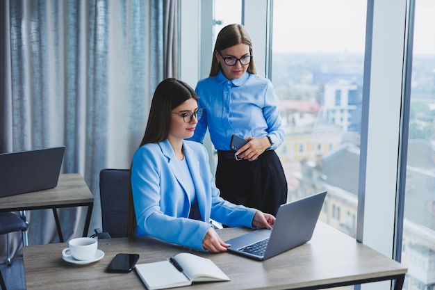 Cheerful employees chatting while working on a laptop discussing work during a coffee break in the office a business woman in glasses listens to a colleague has a pleasant conversation friendships