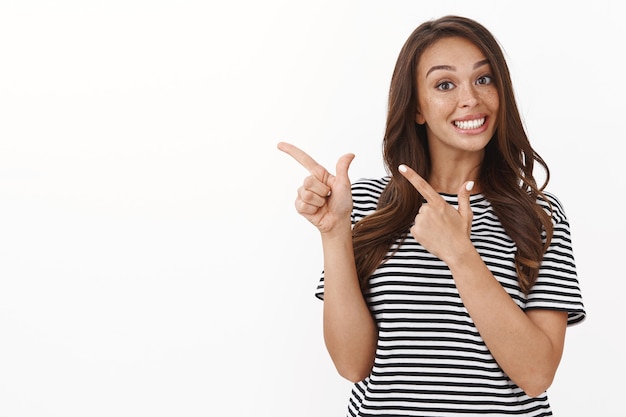 Cheerful enthusiastic young girl suggest link, smiling joyfully tilt head and gazing amused as pointing upper left corner, give personal recommendation, promote product, white wall