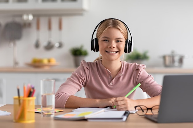 Cheerful european teen girl blonde in headphones study at home at table with laptop in kitchen