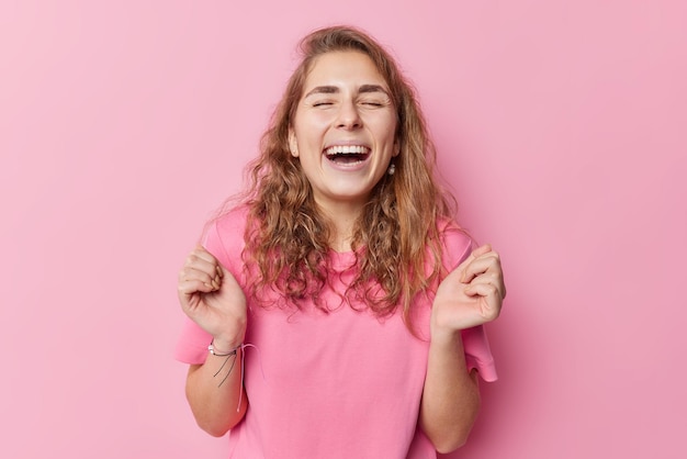 Cheerful European woman shakes arms laughs gladfully keeps eyes closed has upbeat mood wears casual t shirt feels very positive isolated over pink background. Sincere human emotions concept.