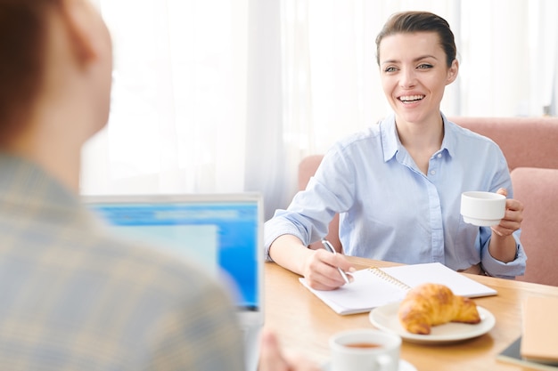 Cheerful excited friendly business lady drinking coffee and writing in notebook while interacting with colleague by lunch