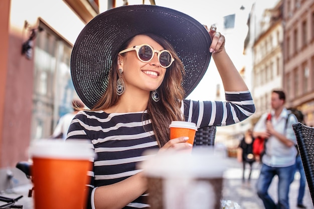 Cheerful fashionable woman wearing sunglasses and stylish hat holding a cup of coffee while sitting in a summer street cafe.