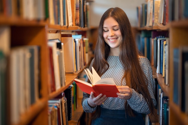 Cheerful female student standing between bookshelves in library of university during break between lesson Happy young woman enjoys reading concept of education und getting knowledge