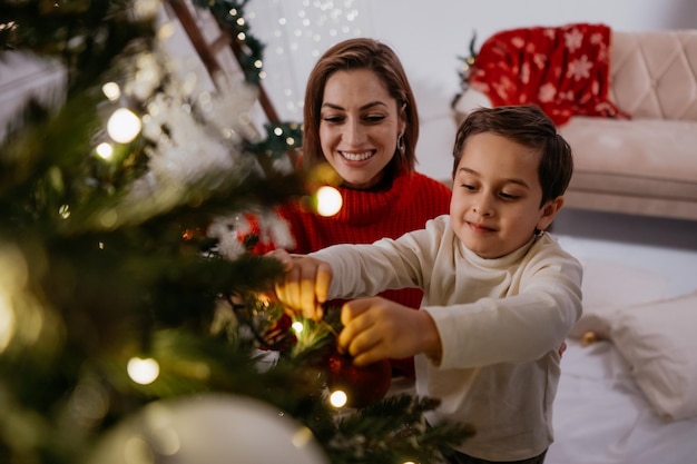 Cheerful festive family of woman with small child son decorating Christmas tree with lights and toys