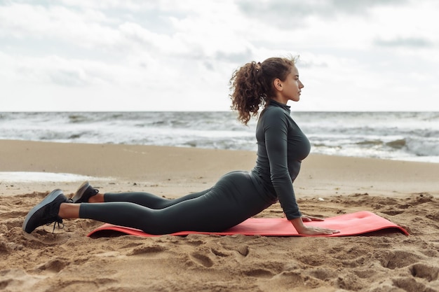 Cheerful fitness woman with curly hair is practicing yoga asana cobra pose on the sand at the beach