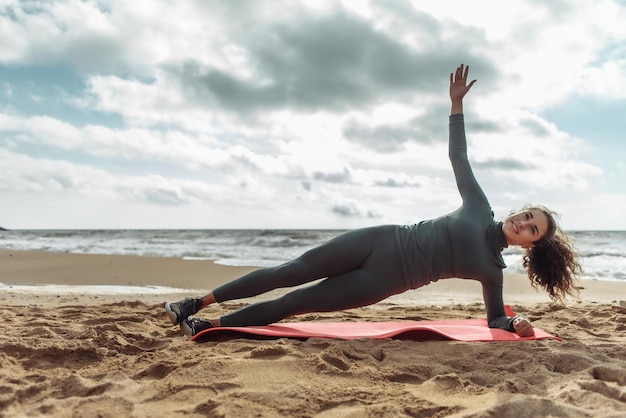 Cheerful fitness woman with curly hair practicing side plank on the sand at the beach