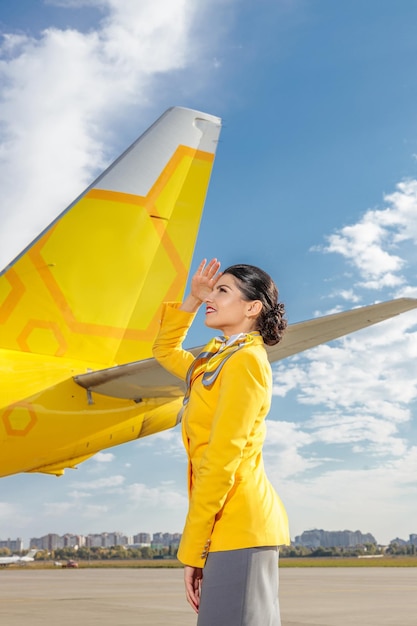 Cheerful flight attendant covering eyes from sun with her hand and smiling while standing near yellow plane at airport