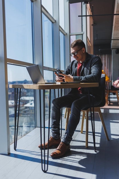 Cheerful freelancer in glasses browsing smartphone and smiling while sitting at table with laptop and notepad in cafe during daytime Working remotely from the office