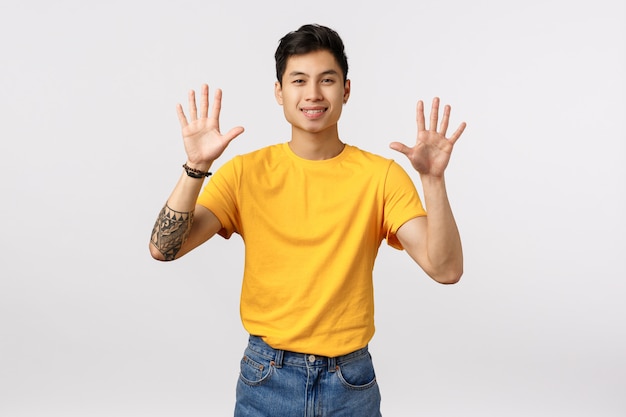 Cheerful, friendly-looking attractive asian man in yellow t-shirt, with tattoos, showing ten fingers, raising arms, order dozen, smiling joyfully, standing white wall upbeat