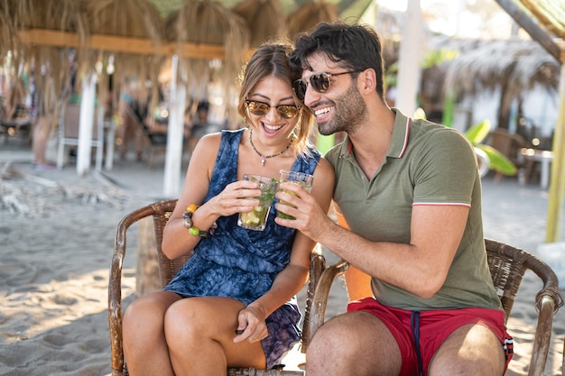 Cheerful friends clinking with mojito cocktails sitting at the beach cafe