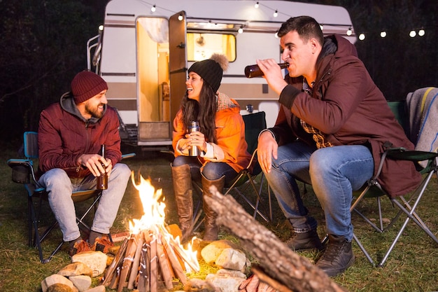 Cheerful friends laughing and drinking beer in a camp site around camp fire. Retro camper van.