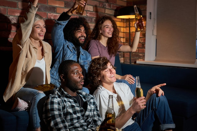 Cheerful friends sitting at home and watching football game on television. Their favorite team is winning, victory concept. Cheering, drinking beer, having rest