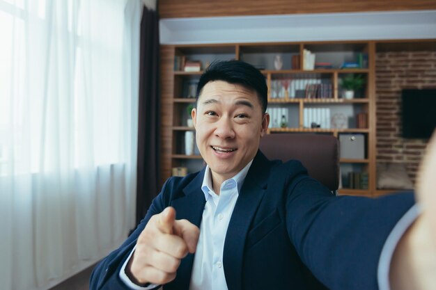 Cheerful and funny Asian businessman looks at the camera of a smartphone talks on a video call a man works in the office waves a congratulatory gesture