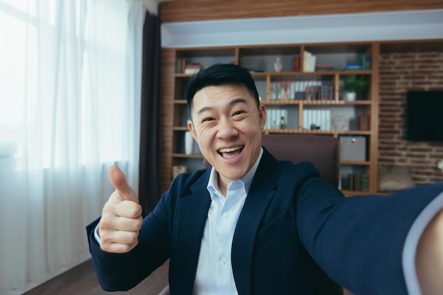 Cheerful and funny Asian businessman looks at the camera of a smartphone talks on a video call a man works in the office waves a congratulatory gesture