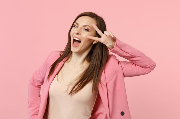 Cheerful funny young woman in jacket keeping mouth wide open, showing victory gesture isolated on pastel pink wall background in studio. People sincere emotions, lifestyle concept. Mock up copy space.