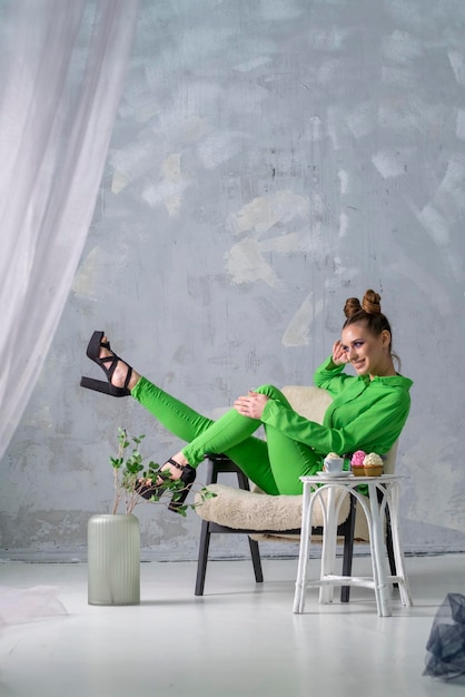 Cheerful girl in green shirt and trousers sits on chair near table with coffee and cupcakes Beautiful woman in grey studio