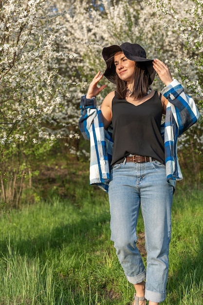 Cheerful girl in a hat among the flowering trees in the spring, in a casual style