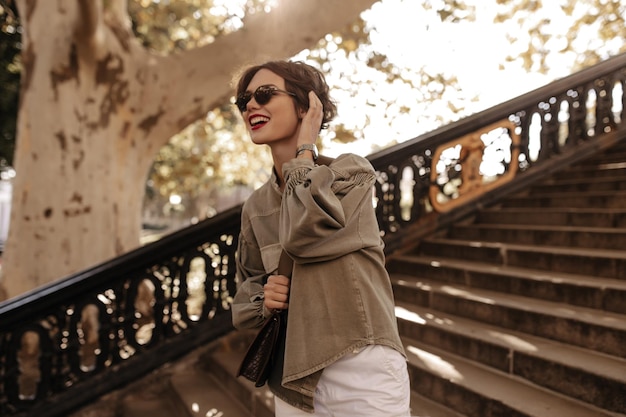 Cheerful girl in olive denim jacket and white pants smiling on backdrop of stairs Joyful lady with handbag and sunglasses poses outside