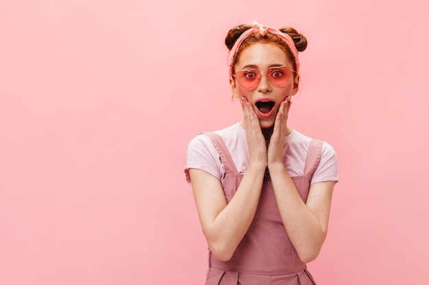 Cheerful girl in pink outfit and sunglasses in joyful surprise looks at camera on isolated background