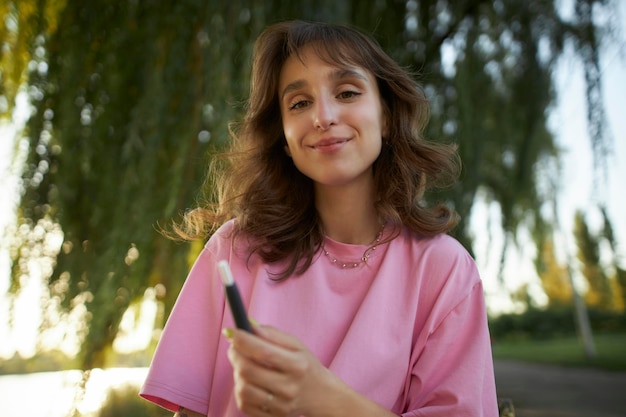 Cheerful girl in a pink t-shirt smile, holding an electronic cigarette, in the park.