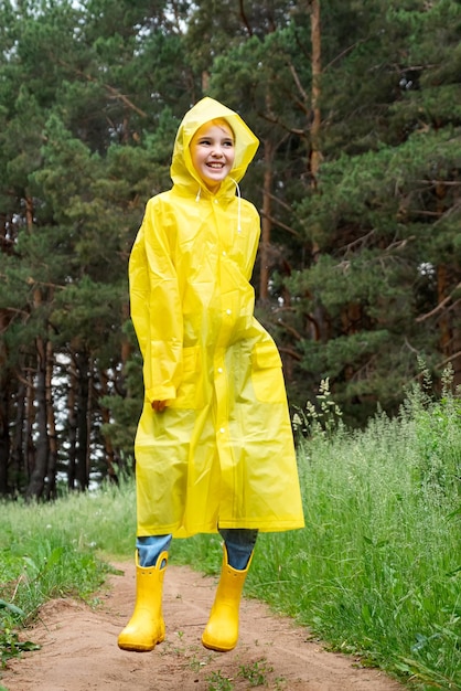 Cheerful girl in rainclothes jumps on path by green forest