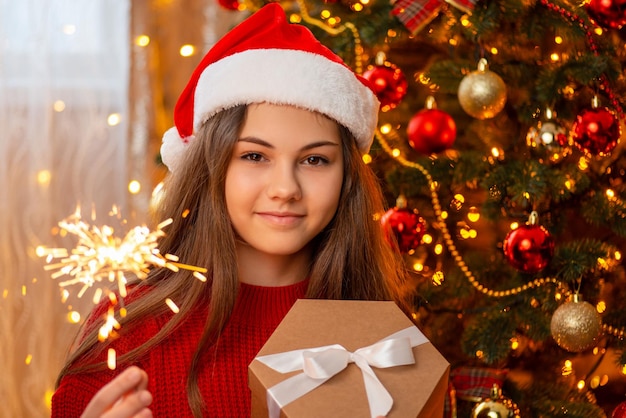 Cheerful girl in santa hat holding present box and burning sparkler